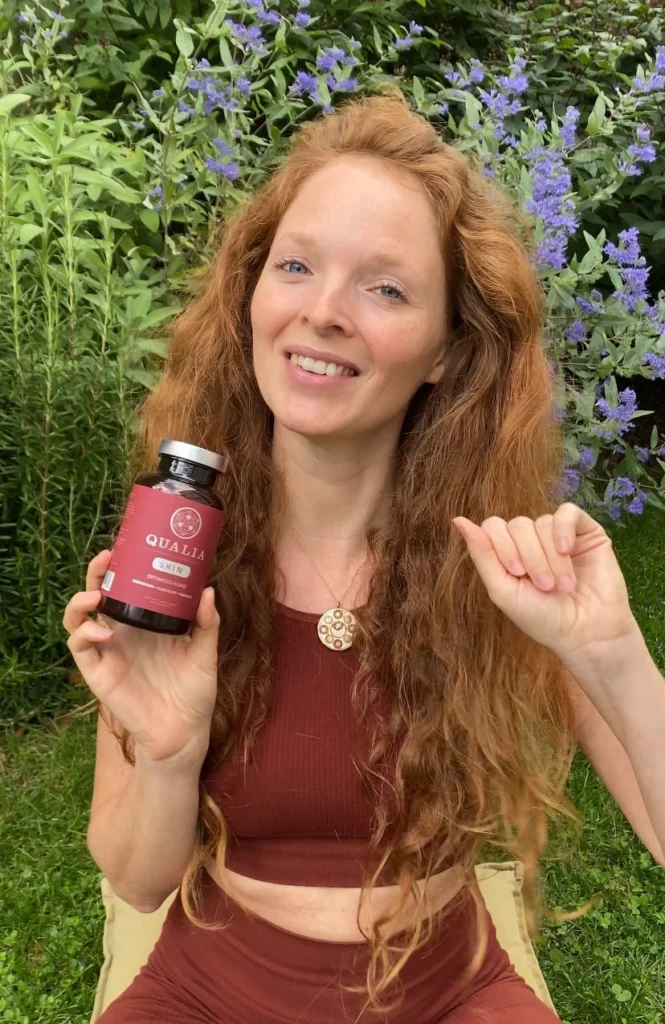 Redhead girl sitting in a garden and holding a bottle of Qualia Skin in her right hand